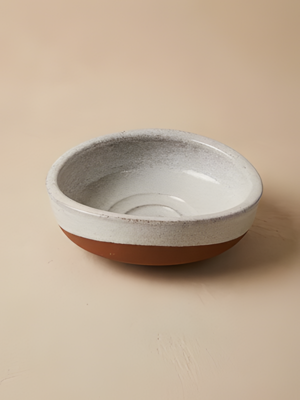 کاسه سفالی دست‌ساز دفورمه با لعاب سفید/خاکستری، بر روی سطحی کرم‌رنگ.
